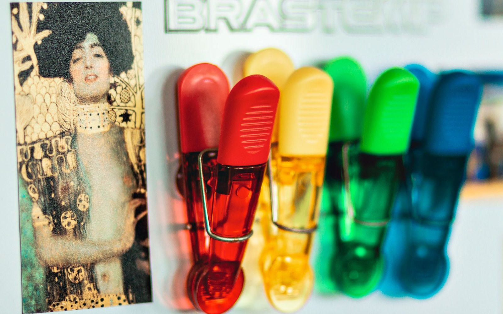 Colorful magnetic clips lined up on a fridge beside a Klimt-style painting magnet