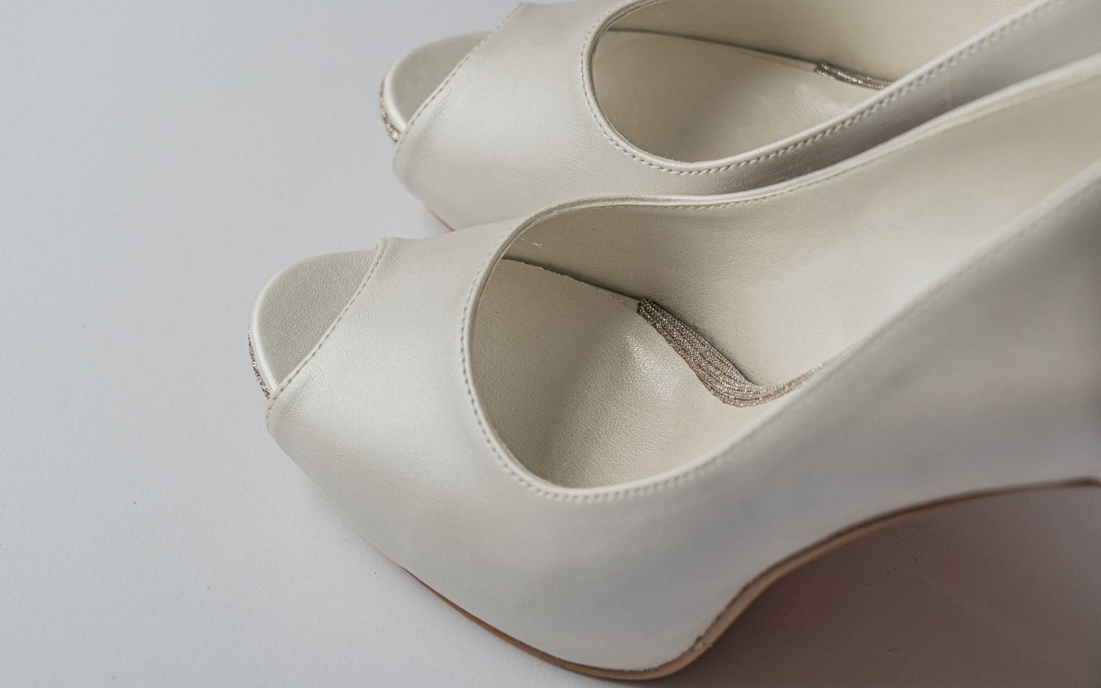 Close-up of white peep-toe wedge shoes with beaded heel detailing on a pale surface