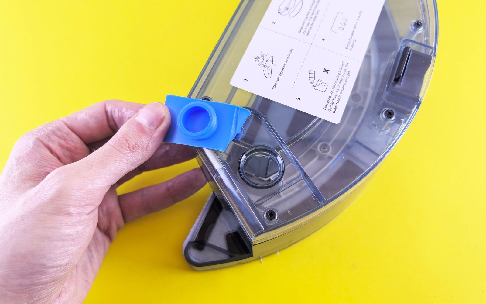 A hand holds a blue plastic cap beside a robot vacuum's clear dustbin lid