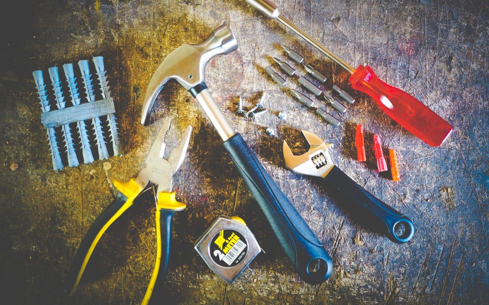 Hammer, pliers, wrench, screwdriver bits and wall anchors arranged on a scratched metal surface