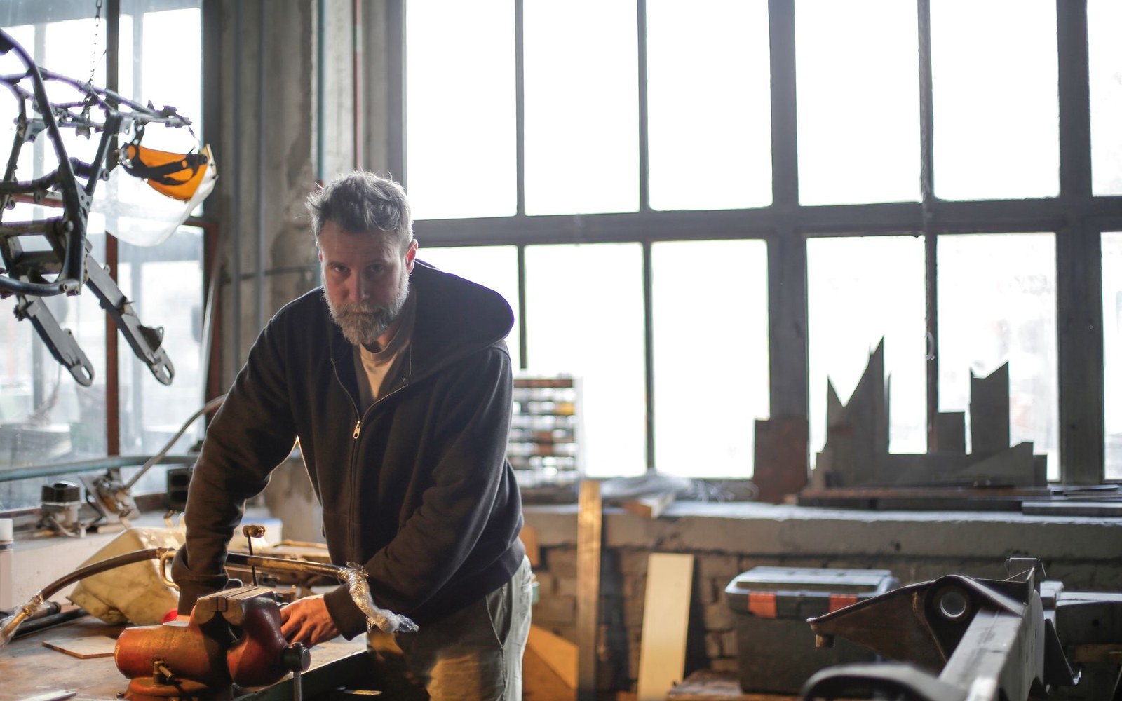 A bearded man stands at a workbench in a workshop filled with tools and metal scraps